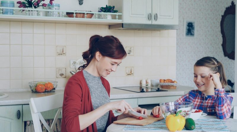 Happy young mother cooking vegetables cutting tomatoes and talking to her cheerful daughter in the kitchen at home. Family, cook, and people concept
